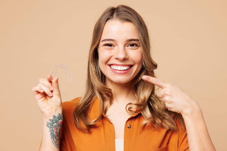 A young woman points to her smile while holding her Invisalign aligner.
